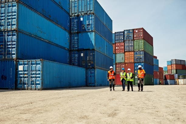 Cargo handlers in hard hats walking between stacks of shipping containers